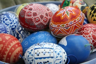 A collection of intricately painted Easter eggs with vibrant and detailed designs. The eggs feature a variety of patterns including floral motifs, geometric shapes, and symmetrical designs, predominantly in red and blue hues.