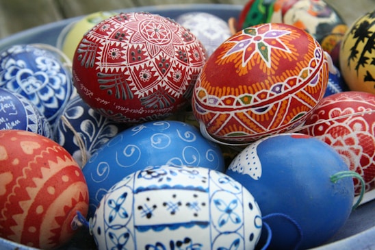 A collection of intricately painted Easter eggs with vibrant and detailed designs. The eggs feature a variety of patterns including floral motifs, geometric shapes, and symmetrical designs, predominantly in red and blue hues.