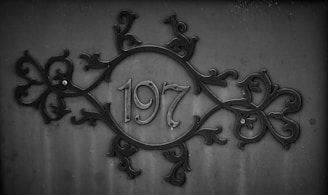 An ornate metal design features the number 197 at its center, surrounded by intricate scrollwork patterns on a dark, textured background. The overall composition suggests a vintage or industrial aesthetic.