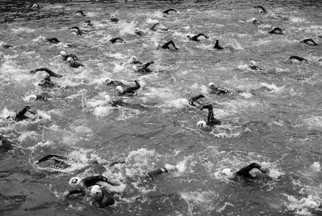 A large group of swimmers wearing caps is engaged in an open water race, creating splashes as they move through the water. The image is in black and white, capturing the dynamic motion of the swimmers against the water's surface.
