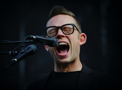A person with glasses enthusiastically singing into a microphone. The mouth is open wide, conveying a sense of energy and passion. The background is dark, highlighting the person's facial expression and the microphone.