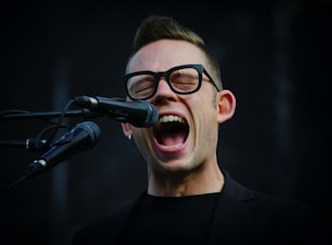 A person with glasses enthusiastically singing into a microphone. The mouth is open wide, conveying a sense of energy and passion. The background is dark, highlighting the person's facial expression and the microphone.