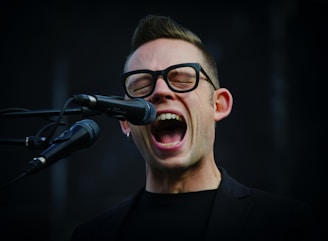 A cheerful bald man singing passionately in front of a microphone with anime posters in the background.