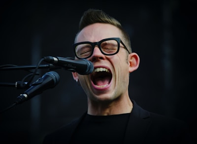 A person with glasses enthusiastically singing into a microphone. The mouth is open wide, conveying a sense of energy and passion. The background is dark, highlighting the person's facial expression and the microphone.
