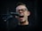 A person with glasses enthusiastically singing into a microphone. The mouth is open wide, conveying a sense of energy and passion. The background is dark, highlighting the person's facial expression and the microphone.