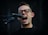 A person with glasses enthusiastically singing into a microphone. The mouth is open wide, conveying a sense of energy and passion. The background is dark, highlighting the person's facial expression and the microphone.