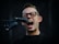 A person with glasses enthusiastically singing into a microphone. The mouth is open wide, conveying a sense of energy and passion. The background is dark, highlighting the person's facial expression and the microphone.