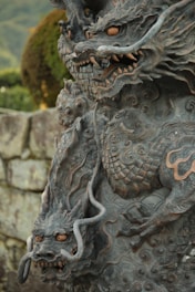 A detailed stone sculpture of a dragon, featuring intricate patterns and fierce facial expressions. The dragon has long, curving horns and sharp, jagged teeth. Organic, swirling motifs adorn its body, adding to the mythical appearance. The background includes a blurred view of greenery and a stone wall.