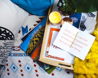 A cozy setup featuring a wooden tray on a patterned blanket with a white pillow. The tray holds a small, lit candle, a succulent plant, and several stacked postcards or booklets. An open notebook is visible with a note defining 'Arete', suggesting a thoughtful or reflective theme.