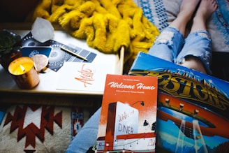 A cozy setting with a person in ripped jeans, lying down with books and a vinyl album on their lap. A candle burns warmly on a tray next to a notebook, pen, and decorative items. The background includes a fluffy yellow blanket, suggesting a relaxed and inviting atmosphere.