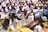 A large gathering of people dressed in white clothing, performing a prayer ritual. Many are wearing floral decorations in their hair and have both hands placed together, raised to their faces in a gesture of prayer or respect.