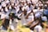 A large gathering of people dressed in white clothing, performing a prayer ritual. Many are wearing floral decorations in their hair and have both hands placed together, raised to their faces in a gesture of prayer or respect.