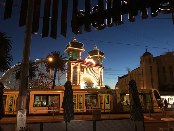 Amusement park entrance illuminated at dusk with decorative lights, including a grand arch and towers. A tram is visible in the foreground, and palm trees are silhouetted against the evening sky.