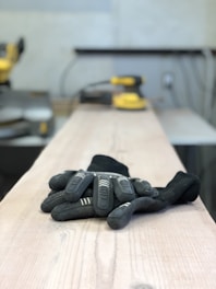A pair of black gloves lies on a wooden surface, likely in a workshop setting. In the background, there is blurred industrial equipment including a sander, giving the impression of a workspace for woodworking or construction.