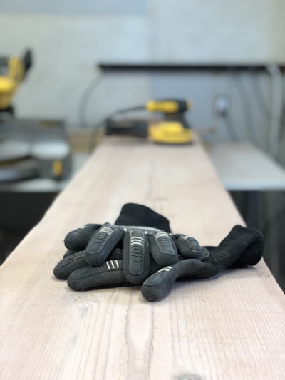 A pair of black gloves lies on a wooden surface, likely in a workshop setting. In the background, there is blurred industrial equipment including a sander, giving the impression of a workspace for woodworking or construction.