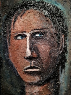 A textured, abstract portrait of a human face with a serious expression. The features are roughly painted and textured, giving a rugged, almost carved appearance. The eyes are deep blue, contrasting against the earthy brown tones of the face. Dark hair is depicted with rough, black strokes.