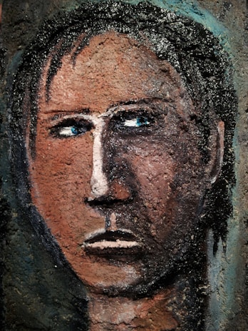 A textured, abstract portrait of a human face with a serious expression. The features are roughly painted and textured, giving a rugged, almost carved appearance. The eyes are deep blue, contrasting against the earthy brown tones of the face. Dark hair is depicted with rough, black strokes.