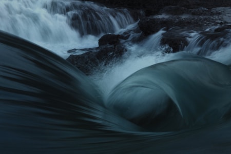 Water flows with strong currents over rocks, swirling and creating smooth, dynamic shapes. The movement is captured with a sense of motion, emphasizing the natural power and fluidity of the water.