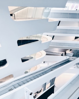 A modern architectural interior with sweeping white curves and multiple levels connected by walkways. The design features extensive use of glass and clean lines, creating a futuristic and open atmosphere. People are seen walking on different levels.