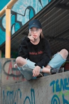 A young person with long hair and a cap sits on a ledge covered in graffiti. They wear a black sweatshirt and light blue ripped jeans. The background includes a metal structure, blue and yellow graffiti, and a yellow railing.
