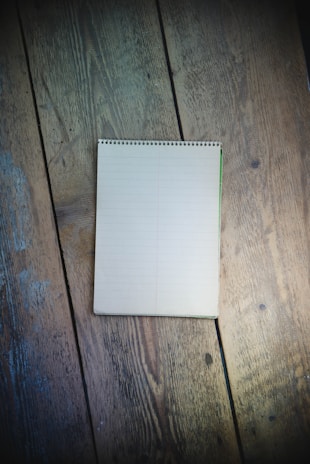 A spiral-bound notebook with lined pages sits open on a rustic wooden surface. The wood shows signs of wear and age, adding texture to the scene. The notebook is blank, suggesting potential for writing or drawing.