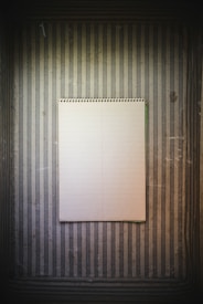A spiral-bound notebook with blank, lined pages placed against a textured background of vertically striped wallpaper in brown and gray tones. The vintage appearance and minimalist setup create a simple yet intriguing composition.