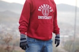 A person is wearing a red Stanford University hoodie paired with blue jeans and gray gloves. The background appears to be an outdoor setting with blurred hills and foliage.
