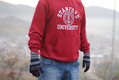 A person is wearing a red Stanford University hoodie paired with blue jeans and gray gloves. The background appears to be an outdoor setting with blurred hills and foliage.