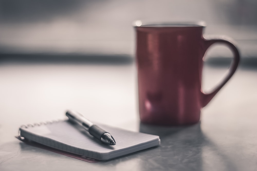 red ceramic mug beside black click pen on white notebook