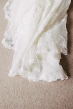 A delicate piece of white lace fabric with intricate floral embroidery patterns is spread out on a textured beige carpet. The lace features scalloped edges and a soft, flowing texture, creating a sense of elegance and sophistication.