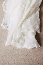 A delicate piece of white lace fabric with intricate floral embroidery patterns is spread out on a textured beige carpet. The lace features scalloped edges and a soft, flowing texture, creating a sense of elegance and sophistication.
