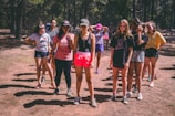 A group of young people is standing in a forest clearing, with trees in the background. They are casually dressed, wearing shorts and t-shirts, and appear to be participating in an outdoor activity or camp. Some are looking down while others face forward.