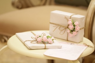 Elegant white gifts adorned with pink rose decorations are tied with rustic twine, resting on a round, cream-colored table next to a piece of stationery.