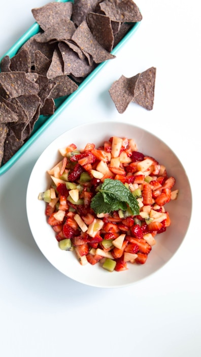 A white bowl filled with a colorful diced fruit salad featuring strawberries, apples, and possibly kiwi, garnished with a fresh mint leaf. Beside the bowl, there is a rectangular green tray containing dark blue tortilla chips, with a couple of chips placed on the white surface nearby.
