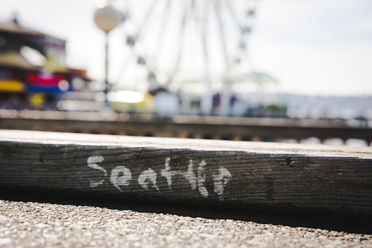 A blurred background with a large wheel structure similar to a Ferris wheel. In the foreground, a wooden surface features the word 'Seattle' in painted white letters. The scene gives an urban setting with a focus on this text, hinting at a city vibe.