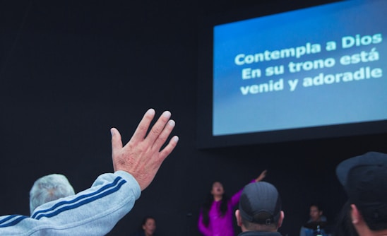 A group of people is gathered in a dark room with a screen displaying religious lyrics in Spanish. One person in the foreground raises a hand in worship, while others are seated or standing towards the back. The setting suggests a religious or worship event.