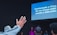A group of people is gathered in a dark room with a screen displaying religious lyrics in Spanish. One person in the foreground raises a hand in worship, while others are seated or standing towards the back. The setting suggests a religious or worship event.