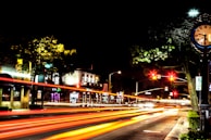 A vibrant city street at night, featuring light trails from passing vehicles creating dynamic lines of red and orange. A vintage clock on a post stands on the right, with its hands pointing around 10:08. The street is lined with trees and buildings, illuminated by streetlights and signs. Traffic lights hang overhead, casting a red glow.