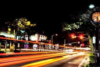 A vibrant city street at night, featuring light trails from passing vehicles creating dynamic lines of red and orange. A vintage clock on a post stands on the right, with its hands pointing around 10:08. The street is lined with trees and buildings, illuminated by streetlights and signs. Traffic lights hang overhead, casting a red glow.