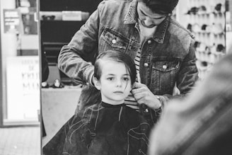 A young child sits in a barbershop chair with a haircut cape draped over, while a barber styles their hair. The barber is wearing a denim jacket and appears focused on their task. The setting is a barbershop with shelves and various items in the background.
