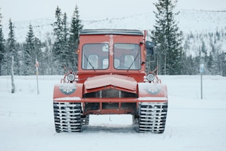 A red snow vehicle with caterpillar tracks stands in a snowy landscape. Tall evergreen trees and snow-covered hills provide a scenic backdrop. The vehicle has large windows and small headlights.