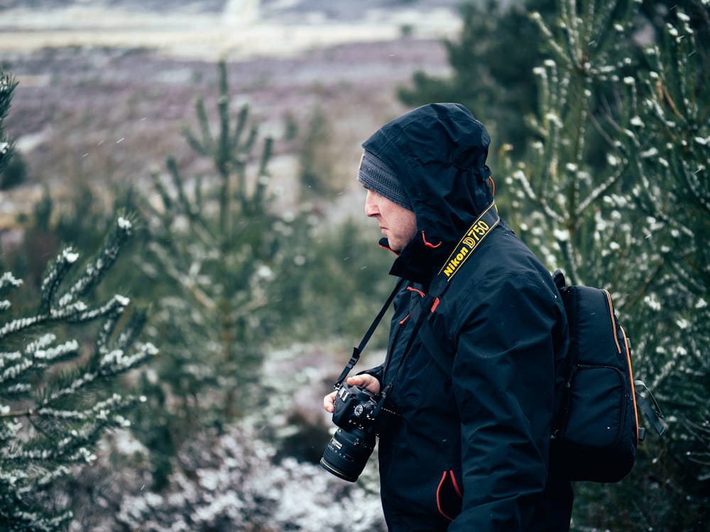 man carrying backpack holding black camera standing beside leafed plants