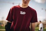 A person is partially visible, wearing a maroon t-shirt with the brand name Tealer on it. They have a black elbow pad on their left arm. The background is blurred, showing greenery and a person in a white shirt walking by.