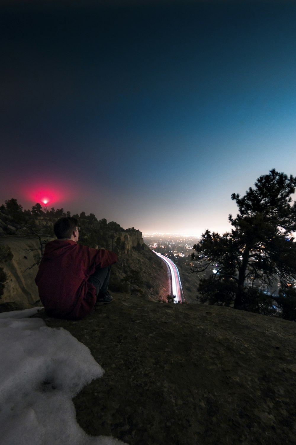 man wearing red hooded jacket sitting viewing mountain and buildings during night time