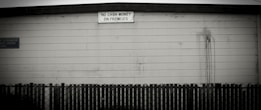 A black and white image depicting the exterior of a building with a weathered wooden facade. A small sign at the top of the wall reads 'NO CASH MONEY ON PREMISES' in capital letters. Below the sign, there is visible streaking from possible water damage. A dark, picket-style fence runs horizontally across the bottom of the image.