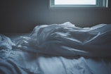 Soft, crumpled white sheets lie on a bed next to a window, suggesting an unmade bed in a dimly lit room.