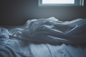 Soft, crumpled white sheets lie on a bed next to a window, suggesting an unmade bed in a dimly lit room.