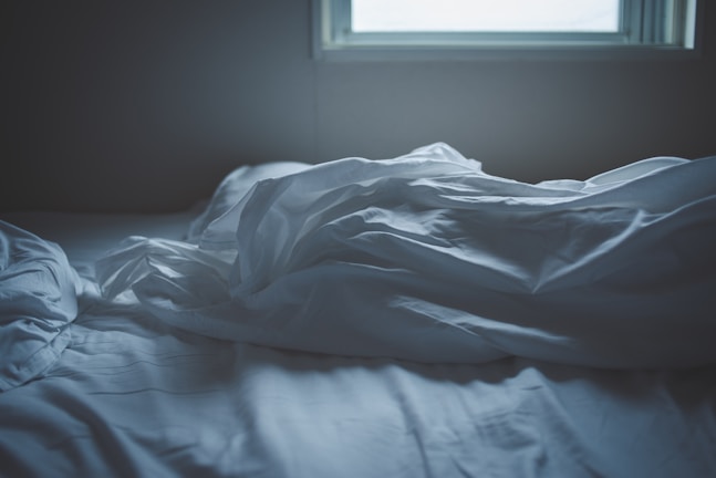 Soft, crumpled white sheets lie on a bed next to a window, suggesting an unmade bed in a dimly lit room.