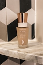A bottle of Charlotte Tilbury Magic Foundation is placed on a marble surface. The bottle features a rose gold cap and contains SPF 15 sunscreen. The background displays a pattern of black, white, and grey geometric tiles.