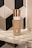 A bottle of Charlotte Tilbury Magic Foundation is placed on a marble surface. The bottle features a rose gold cap and contains SPF 15 sunscreen. The background displays a pattern of black, white, and grey geometric tiles.